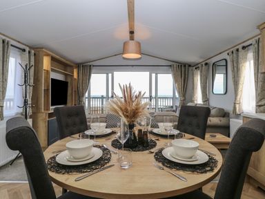 A dining area with a round wooden table set with bowls plates glasses and cutlery in a living room with sofas and a TV at Dorchester in Broomholm near Mundesley