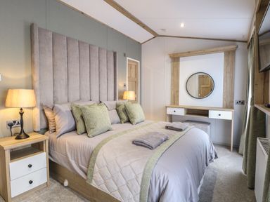 A bedroom with a bed bedside tables lamps a dressing table with a round mirror and a TV on the wall at Dorchester in Broomholm near Mundesley