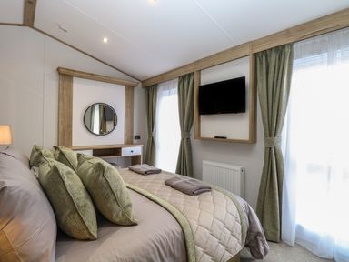 A bedroom with a bed four green pillows a wooden framed wall mirror a mounted television and windows with green curtains at Dorchester in Broomholm near Mundesley