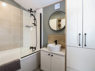 A bathroom with a bathtub and shower fixture a round mirror and a sink with cabinet at Dorchester in Broomholm near Mundesley