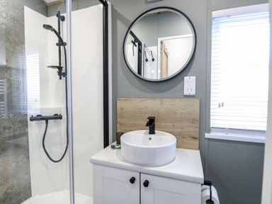A bathroom with a shower enclosure a round mirror a round sink and a window with blinds at Dorchester in Broomholm near Mundesley