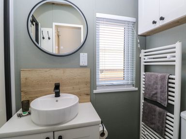A bathroom with a white sink and black faucet a round mirror a window with blinds and a white towel rack with two towels at Dorchester in Broomholm near Mundesley