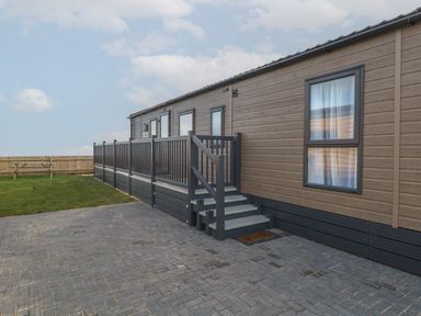 Exterior view of a brown mobile home with stairs and railing on a paved driveway and grass area with a wooden fence and picnic bench at Dorchester in Broomholm near Mundesley