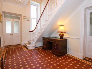 An entryway with a wooden desk and lamp under a staircase with white balusters and a patterned tile floor at Fir Grove in Menai Bridge