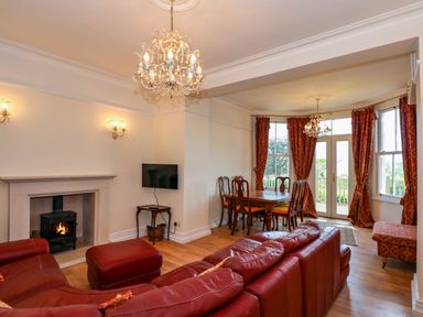 A living room with a red leather sofa a fireplace a dining table with chairs and red curtains at Fir Grove in Menai Bridge