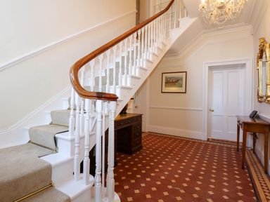 A hallway with a staircase carpeted in grey and wooden handrail a wooden desk and table framed pictures and a chandelier at Fir Grove in Menai Bridge