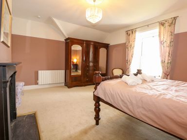 A bedroom with a wooden bed frame bed with towels on it a wooden wardrobe a chair window with floral curtains and a fireplace at Fir Grove in Menai Bridge