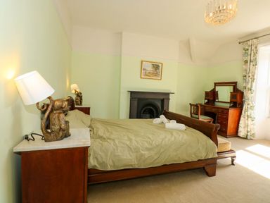 A bedroom with a bed with beige bedding two bedside tables with monkey lamps a fireplace a wooden dressing table with a chair and floral curtains at Fir Grove in Menai Bridge