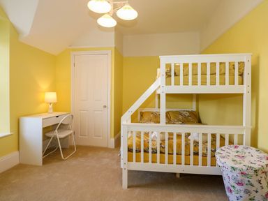 A bedroom with yellow walls a white bunk bed a white desk with a chair and a lamp and a floral storage bin at Fir Grove in Menai Bridge