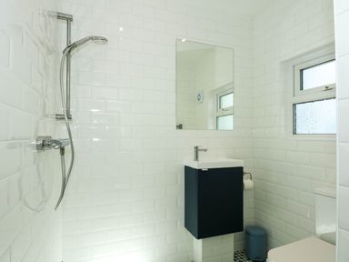 A bathroom with a shower, a small sink with cabinet, a mirror, a window, a trash bin, and a toilet at Fir Grove in Menai Bridge