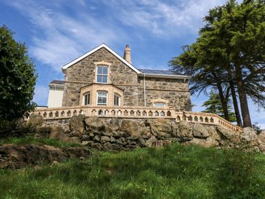 A stone house with brick accents and a balcony above a rocky slope surrounded by grass and trees at Fir Grove in Menai Bridge