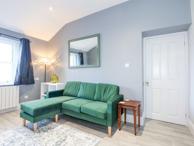 A living room with a green sofa a white door a wooden side table a white shelf with a green vase a floor lamp and a mirror on a grey wall at The Fir Grove Annex in Menai Bridge