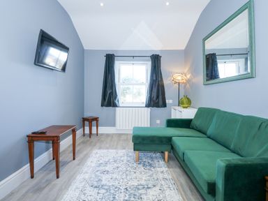 A living room with a green sofa a wall-mounted tv a window with black curtains and a patterned rug at The Fir Grove Annex in Menai Bridge