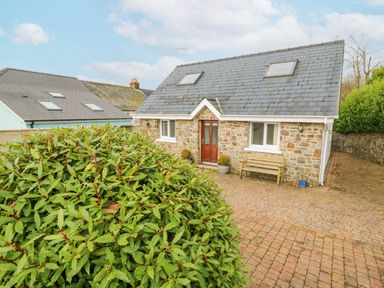 A stone house with a slate roof two windows and a wooden bench on a brick driveway with a green bush in front at Trevone in Goodwick
