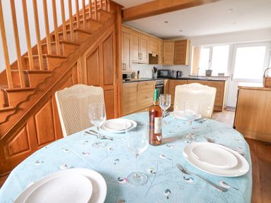 A kitchen with wooden cabinets and stairs in the background and a dining table set with plates glasses and a bottle of wine with a blue tablecloth at Trevone in Goodwick