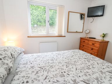 A bedroom with a bed with patterned bedding a wooden dresser a wall mirror a wall-mounted tv and a window at Trevone in Goodwick