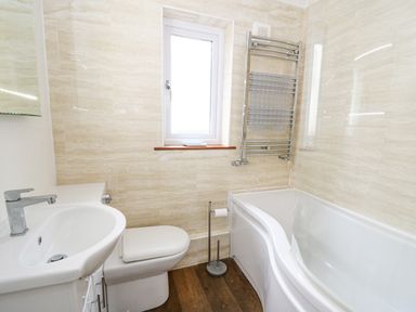 A bathroom with a sink, toilet, bathtub with glass shower screen, wall-mounted towel radiator and small window at Trevone in Goodwick