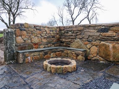 An outdoor stone seating area with a fire pit and a stone wall behind near trees at Ynys Uchaf Near Benllech