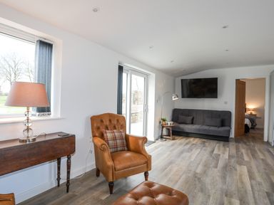 A living room with a brown armchair, a black sofa, a wall-mounted TV, and a wooden table with a lamp at Ynys Uchaf Near Benllech