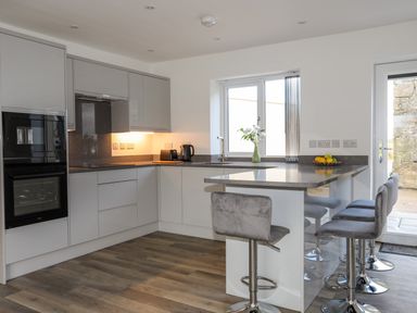 A modern kitchen with gray cabinets an island with gray bar stools and wooden flooring at Ynys Uchaf Near Benllech