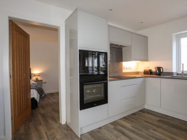 A kitchen with white cabinets black built in oven and microwave wooden floor and an open door to a bedroom at Ynys Uchaf Near Benllech