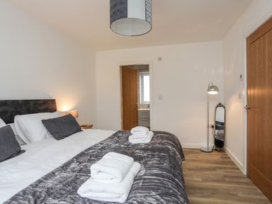 A bedroom with a bed with gray and white bedding towels on the bed a bedside table with a lamp a floor lamp a mirror and two wooden doors at Ynys Uchaf Near Benllech