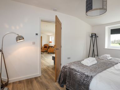 A bedroom with a bed covered with a grey blanket and folded towels next to a window and an open door showing a living area with leather chairs at Ynys Uchaf Near Benllech