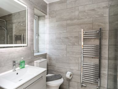 A bathroom with a sink, illuminated mirror, toilet, towel on toilet seat, toilet paper holder, window, and heated towel rail at Ynys Uchaf Near Benllech