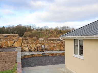 An outdoor patio area with two chairs and a table next to a stone wall and a beige building with a metal roof at Ynys Uchaf Near Benllech