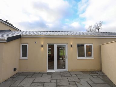 An exterior view of a beige building with a metal roof two windows and a glass door opening to a paved patio at Ynys Uchaf Near Benllech