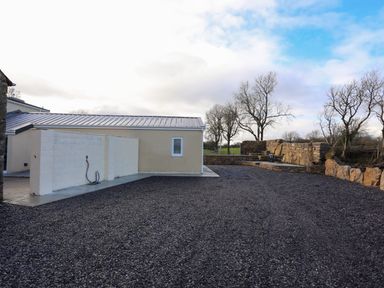 An outdoor area with a beige building a white wall garden hose and stone seating area with chairs near leafless trees at Ynys Uchaf Near Benllech