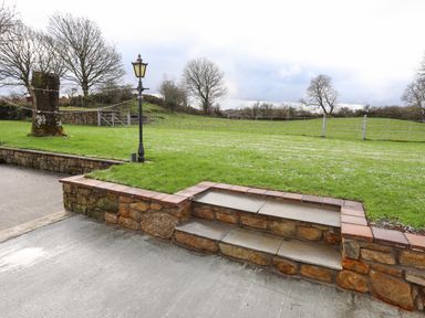 A stone patio with steps leading to a lawn with a black lamp post and metal gates in the background at Ynys Uchaf Near Benllech