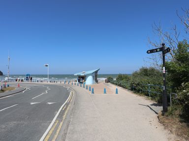 A coastal road next to a small blue building near the sea with a flagpole and a signpost surrounded by bushes near Benllech
