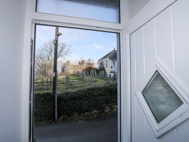 A view through an open white door showing a graveyard with headstones a tree a utility pole and a stone castle beyond at Gwelfor in Harlech
