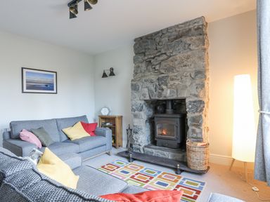 A living room with a stone fireplace a wood stove two grey sofas with colored cushions a small wooden bookshelf a tall floor lamp and a colorful rug at Gwelfor in Harlech