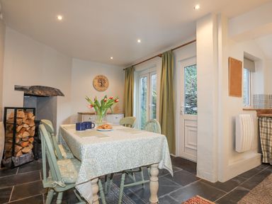A dining area with a wooden table, green chairs, a vase of tulips, a clock on the wall, a wood storage rack, and green curtains over glass doors at Gwelfor in Harlech