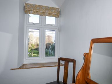 A white room with a wooden chair and a mirror near a window overlooking a graveyard and a stone castle at Gwelfor in Harlech