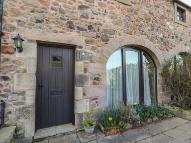 A stone exterior wall with a dark wooden door a lamp and an arched window with plants outside at Primula Patch Akeld near Wooler