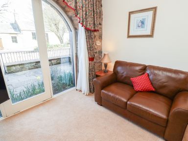 A brown leather sofa with a red cushion near a window with floral curtains and a side table with a lamp at Primula Patch in Akeld near Wooler