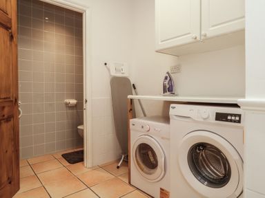 A laundry room with a washing machine dryer ironing board iron and open door showing tiled bathroom at Hilltop Cottage in Paignton
