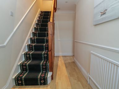 A staircase with a patterned carpet runner and a wooden banister in a hallway with light-colored walls and wooden flooring at Hilltop Cottage in Paignton