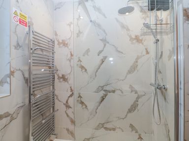 A shower area with marble tile walls a chrome towel radiator and a rainfall showerhead at Hilltop Cottage in Paignton