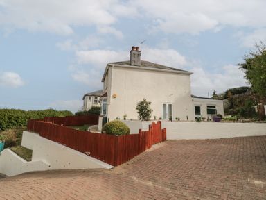 A paved driveway leading to a white two-story house with red wooden fencing and small garden plants at Hilltop Cottage in Paignton