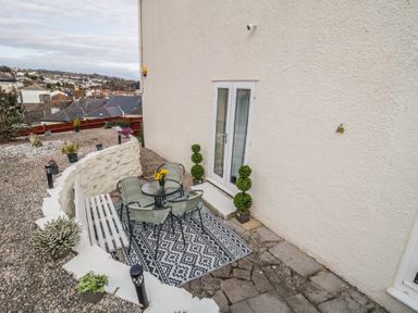 An outdoor patio area with a round glass table and four chairs on a patterned rug next to a white bench and potted plants at Hilltop Cottage in Paignton