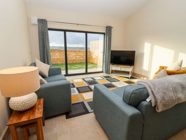 A living room with two blue sofas a lamp on a wooden side table a television on a white stand and sliding glass doors leading outside at Bears Court Barn 1 Little Rissington near Bourton-On-The-Water