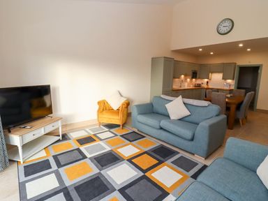 A living room with a patterned rug blue sofas a yellow armchair and a television next to a kitchen with dining table and chairs at Bears Court Barn 1 in Little Rissington near Bourton-On-The-Water