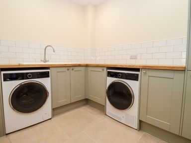 A utility room with a washing machine dryer under a wooden countertop with green cabinets and white tiled backsplash at Bears Court Barn 1 in Little Rissington near Bourton-On-The-Water