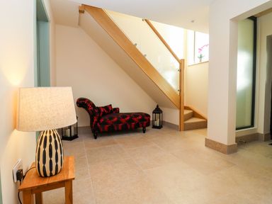 An entryway with a patterned chaise lounge under a wooden staircase and a lamp on a small wooden table at Bears Court Barn 1 Little Rissington near Bourton-On-The-Water