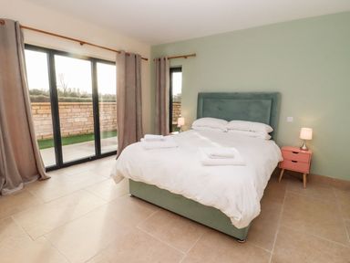 A bedroom with a double bed white bedding bedside tables lamps stone tile floor and sliding glass doors with curtains at Bears Court Barn 1 Little Rissington near Bourton-On-The-Water