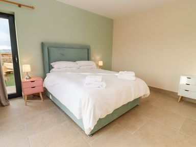 A bedroom with a double bed with a green upholstered headboard white bedding towels on the bed two bedside tables with lamps and a large glass door at Bears Court Barn 1 Little Rissington near Bourton-On-The-Water